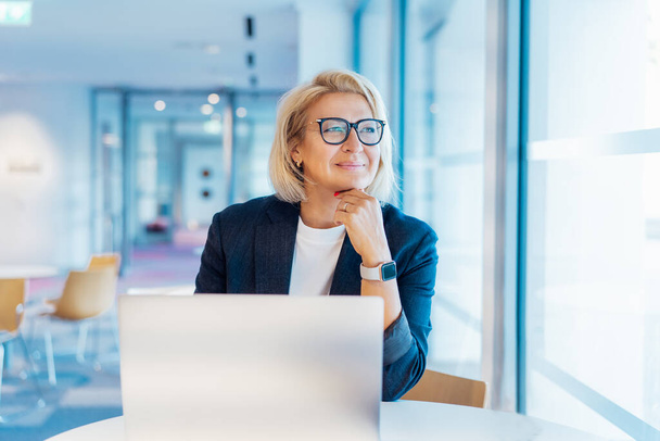 50s confident mature businesswoman dreamy looking at window, middle-aged experienced senior female professional working on laptop in open space office. Female entrepreneur working remotely. - 写真・画像