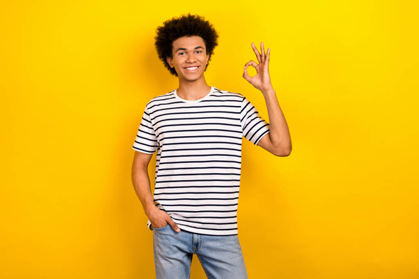 Photo funny student guy good mood showing okey symbol approval wear striped t-shirt jeans isolated yellow color background. - Foto, imagen