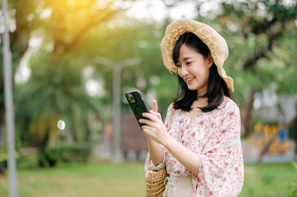 Portrait of young asian woman traveler with weaving hat and basket using mobile phone on green park nature background. Journey trip lifestyle, world travel explorer or Asia summer tourism concept. - 写真・画像