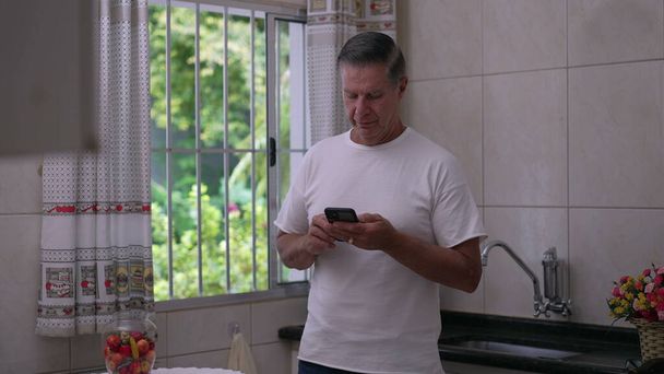 Un uomo di mezza età che guarda lo schermo del dispositivo smartphone mentre si trova nella cucina di casa vicino alla finestra. Uomo anni '50 persona che utilizza la tecnologia moderna in stile di vita casual domestico - Foto, immagini