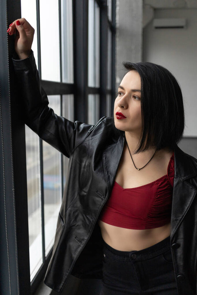 A girl with black hair, red lipstick and a leather jacket stands near a window - Foto, Imagen