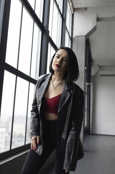 A girl with black hair, red lipstick and a leather jacket stands near a window - Foto, immagini