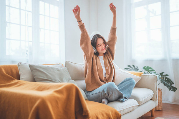 Щаслива азіатська жінка слухає музику з мобільного телефону, сидячи на дивані вдома, усміхнена дівчина розслабляється з навушниками вранці, час розслабитися. копіювання простору
. - Фото, зображення