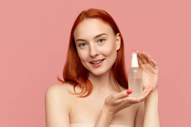 Fashion model showing bottle of serum and smiling over pink studio background. Facial cosmetics product, masks. Concept of natural beauty, skin care, cosmetology, health, ad - Photo, image