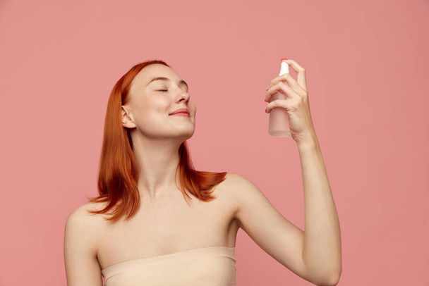 Young happy girl with red hair using moisturising spray on face with closed eyes over pink studio background. Facial cosmetics product, masks. Concept of natural beauty, skin care, cosmetology, ad - Photo, Image