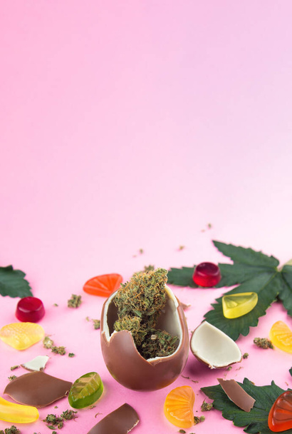 Chocolate egg with dry buds of medical marijuana inside on a pink background.  Surrounded by cannabis leaves, gummies and chocolate chips - Photo, Image