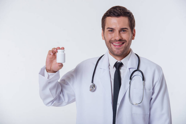 Handsome doctor in white coat is holding a bottle of medicine, looking at camera and smiling, isolated on white background - Fotografie, Obrázek
