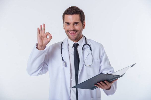Портрет красивого лікаря в білому пальто, що тримає папку, показує знак ОК, дивлячись на камеру і посміхаючись, ізольовані на білому тлі
 - Фото, зображення