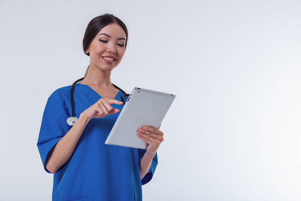 Portrait of beautiful Asian female doctor in blue medical wear using a tablet and smiling, isolated on white background - Fotó, kép