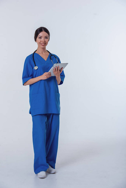 Full length portrait of beautiful Asian female doctor in blue medical wear holding a laptop, looking at camera and smiling, isolated on white background - Foto, Bild