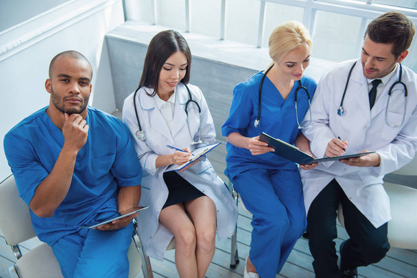 Group of medical doctors of different nationalities and genders with folders and tablet is examining documents and smiling, sitting in office. One is looking at camera - Fotografie, Obrázek