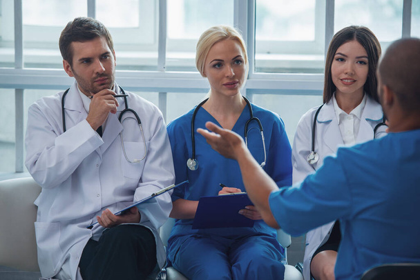 Ärzte verschiedener Nationalitäten und Geschlechter machen sich Notizen, während sie sich im Büro den Bericht eines anderen Arztes anhören - Foto, Bild