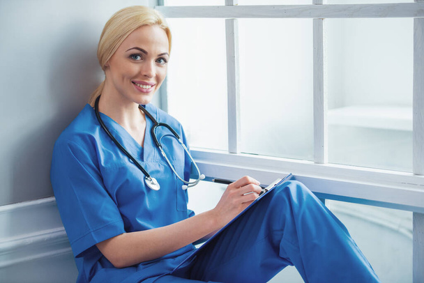 Mooie blonde vrouwelijke arts in blauwe medische slijtage is het maken van notities, camera kijken en lacht, zitten in de buurt van het venster - Foto, afbeelding