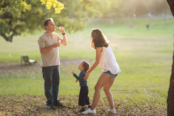 Onnellinen valkoinen perhe viettää aikaa puistossa isä puhaltaa saippuakuplia. Keskimmäinen laukaus - Valokuva, kuva