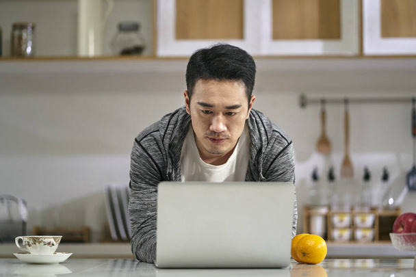 молодой азиатский бизнесмен, работающий на кухне дома с помощью ноутбука - Фото, изображение
