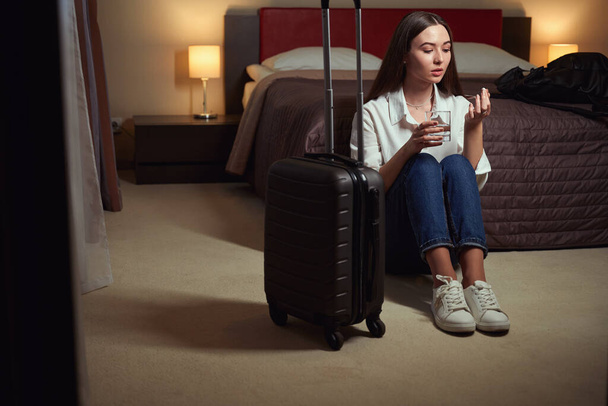 Krásná žena drží sklenici vody a tablet, zatímco sedí na podlaze v blízkosti postele v hotelovém pokoji - Fotografie, Obrázek