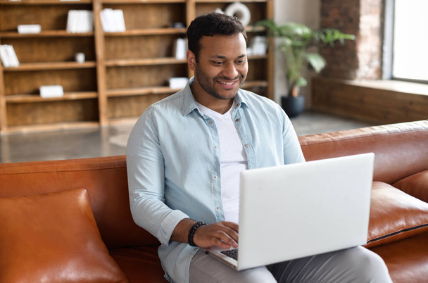 Cheerful happy handsome indian guy in casual wear using laptop sitting on the leather couch with bookshelves on the background, dark-haired hispanic man web surfing, spends leisure in social network - Photo, Image