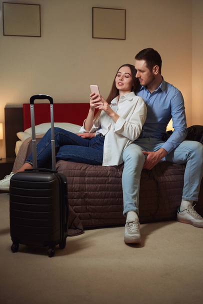 Junge schöne Dame zeigt ihrem Freund ihr Handy, während sie im Hotelzimmer auf dem Bett sitzt - Foto, Bild