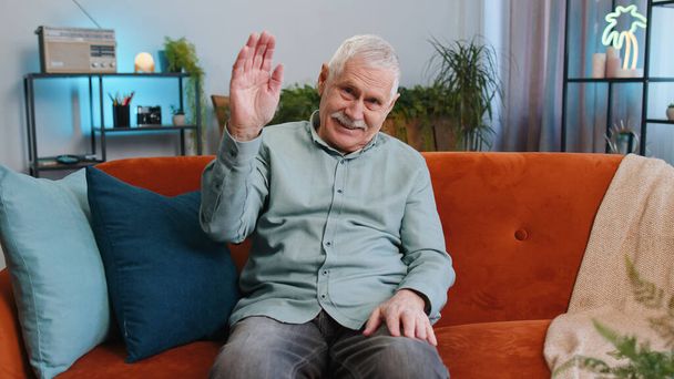 Hello. Grandfather senior man smiling friendly at camera, waving hands gesturing hello hi, greeting or goodbye, welcoming with hospitable expression at home. Elderly guy sitting on sofa in living room - Photo, image