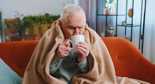 Enfermo abuelo mayor hombre en cuadros temblando de frío en el sofá beber té caliente en apartamento sin calefacción debido a la deuda. Enfermo enfermo jubilado malsano sensación de malestar tratar de calentar - Foto, Imagen
