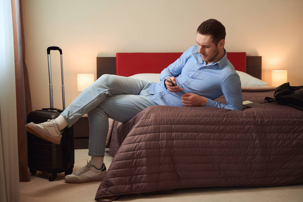 Νεαρός με γενειάδα ακουμπισμένος στο χέρι του ξαπλωμένος στο μεγάλο κρεβάτι με τη βαλίτσα δίπλα του στο δωμάτιο του ξενοδοχείου - Φωτογραφία, εικόνα