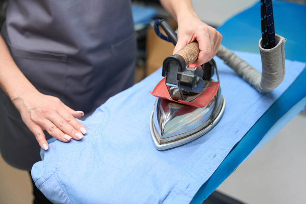 Close-up woman using hot iron to remove wrinkles from shirt, professional electric appliance at dry-cleaning office - Photo, image