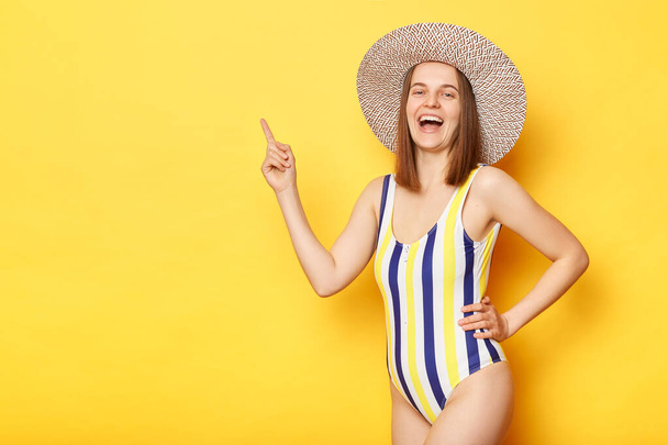 Overjoyed woman wearing striped one-piece swimsuit and straw hat posing isolated over yellow background laughing pointing at advertisement area space for your text. - Zdjęcie, obraz