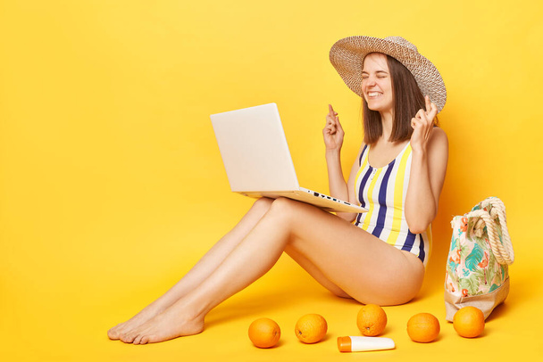 Hopeful woman wearing striped one-piece swimsuit and straw hat posing isolated over yellow background booking tickets for vacation online praying to have free dares for departure. - Valokuva, kuva