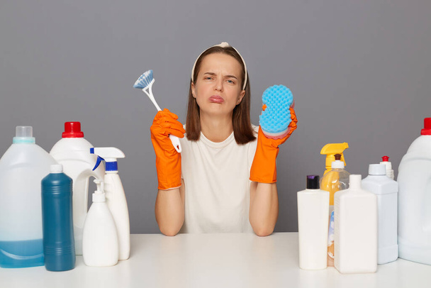 Sad upset unhappy woman posing at workplace with cleaning detergents holding sponge and brush crying looking at camera with pout lips isolated over gray background. - Foto, Bild
