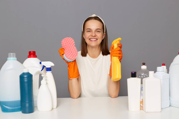 Attractive smiling woman housewife posing at workplace with cleaning detergents isolated over gray background looking at camera holding sponge and cleaning spray. - Photo, Image
