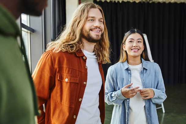 cheerful and trendy long haired man smiling near young asian woman and african american colleague on blurred foreground in modern office, successful collaboration, creative startup - Fotoğraf, Görsel