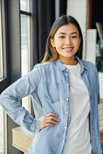 junge attraktive asiatische Geschäftsfrau in blauem Jeanshemd, mit brünetten Haaren und strahlendem Lächeln, die mit der Hand auf der Hüfte steht und in die Kamera im modernen Büro blickt, positive Emotion - Foto, Bild