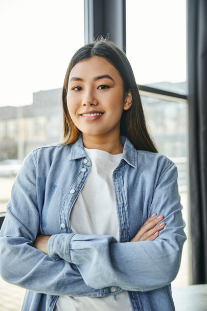 стильная и радостная азиатская брюнетка в джинсовой рубашке, скрещивающая руки и смотрящая в камеру в современном офисе, успешная молодежь, профессиональное развитие, современный образ жизни - Фото, изображение