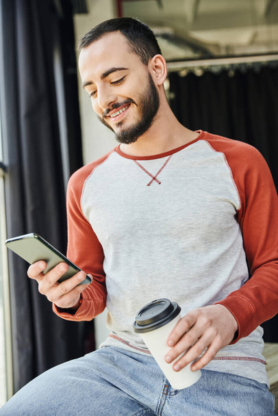 charismatic entrepreneur, bearded and stylish, holding coffee to go in paper cup and browsing internet on smartphone, young and career oriented businessman in modern office - 写真・画像