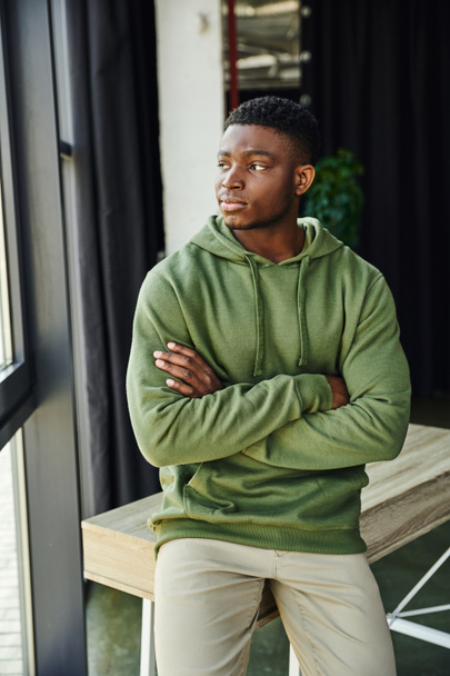 töprengő afro-amerikai férfi zöld elegáns kapucnis ül az asztalon összecsukott karokkal, és félrenéz a modern irodában, fiatal üzletember, szakmai fejlődés  - Fotó, kép
