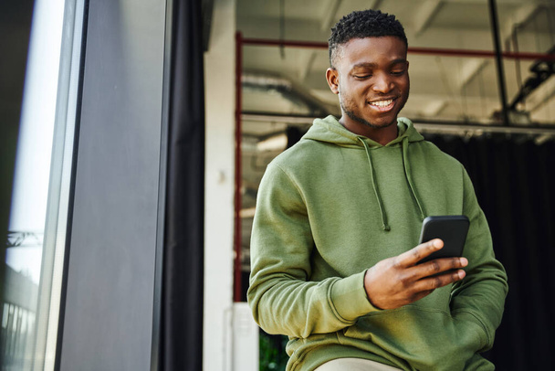 jovem e alegre homem afro-americano segurando mão no bolso de capuz verde e navegar na internet no telefone móvel no escritório moderno, empresário de sucesso, emoção positiva  - Foto, Imagem