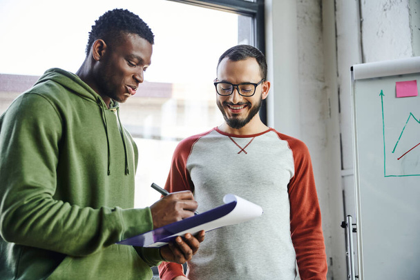 young african american businessman in green hoodie writing on clipboard near smiling bearded colleague in eyeglasses and flip chart with graphs in modern office, creative youth  - Foto, Imagem