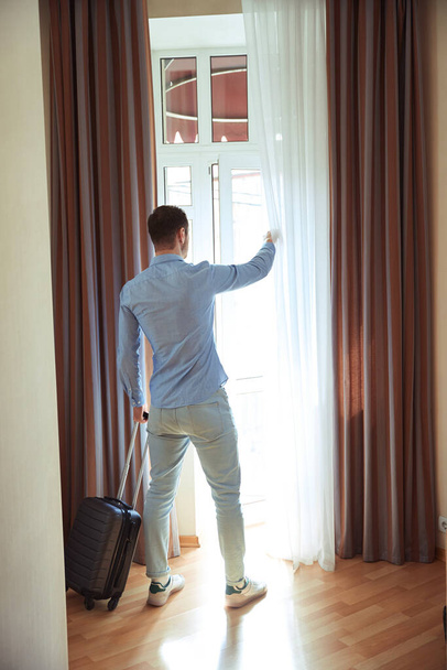 Young man opens a curtained window while standing with a suitcase in a hotel room. View from the back - Φωτογραφία, εικόνα