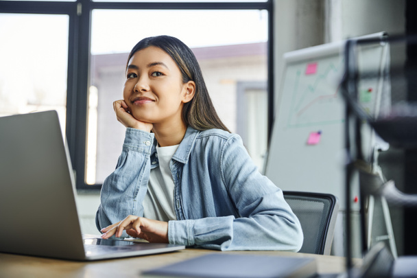 radosna azjatycka bizneswoman w niebieskim dżinsowej koszuli, z brunetka włosy siedzi obok laptopa, trzymając się za rękę blisko twarzy i patrząc daleko w biurze, młody odnoszący sukcesy przedsiębiorca  - Zdjęcie, obraz