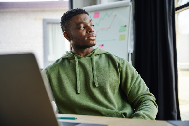 jovem empresário afro-americano com capuz verde sentado e olhando para longe ao planejar a inicialização perto de flip chart com análise de negócios no escritório moderno, desenvolvimento profissional  - Foto, Imagem