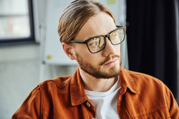 portrait of thoughtful and bearded businessman in eyeglasses and trendy shirt looking away in contemporary office, thinking and planning startup project, blurred background - Foto, Imagem
