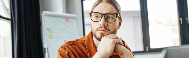 portrait of serious and thoughtful bearded businessman in eyeglasses and trendy shirt holding hands under chin and looking away near flip chart on blurred background in office, banner - Photo, Image