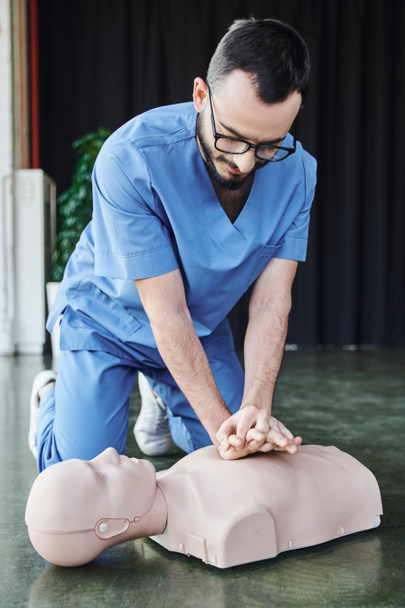 junger medizinischer Instruktor mit Brille und Uniform beim Durchführen von Brustkompressionen an der CPR-Schaufensterpuppe auf dem Boden im Schulungsraum, effektive lebensrettende Fähigkeiten und Notfallvorsorgekonzept - Foto, Bild