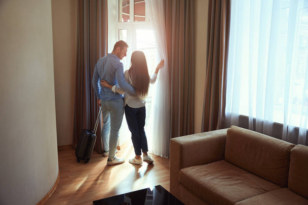 Loving young couple hugging stands with a suitcase in a hotel room and looks out the window - Photo, Image
