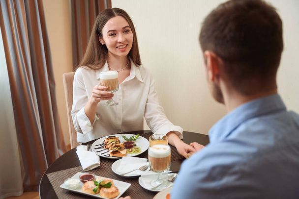 Młody facet i kobieta jedzą śniadanie siedząc przy stole w jasnym hotelu. Zameldowanie - Zdjęcie, obraz