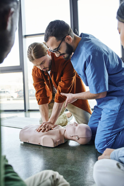 Erste-Hilfe-Seminar, professionelle Sanitäter helfen jungen Mann bei Brustkompressionen an CPR-Schaufensterpuppen in der Nähe multiethnischer Teilnehmer, effektive lebensrettende Fähigkeiten und Techniken Konzept - Foto, Bild