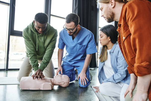 młodzież wieloetniczna i instruktor medycyny patrzący na Afrykańczyka, który uciska klatkę piersiową na manekinach resuscytacyjnych, resuscytacji serca, seminarium szkoleniowe z pierwszej pomocy - Zdjęcie, obraz