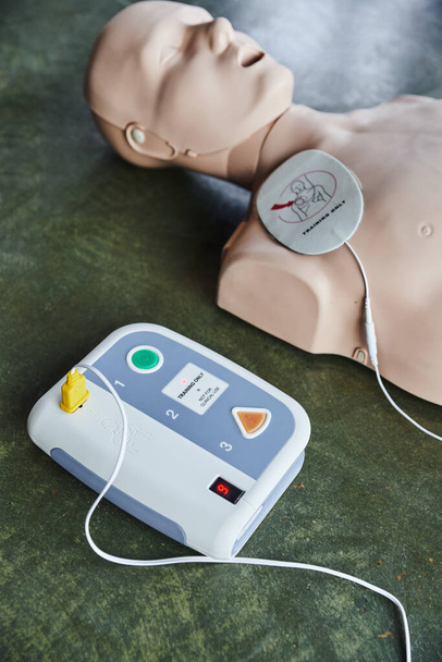 hooghoekaanzicht van een geautomatiseerde externe defibrillator in de buurt van een cardiopulmonale reanimatiepop van de vloer in de trainingsruimte, medische apparatuur voor EHBO-training en ontwikkeling van vaardigheden - Foto, afbeelding