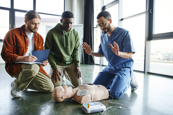 νεαρός τραυματιοφορέας που εξηγεί τεχνικές καρδιακής ανάνηψης σε διαφυλετικούς συμμετέχοντες κοντά στο ανδρείκελο CPR με αυτόματο εξωτερικό απινιδωτή, αποτελεσματικές δεξιότητες και τεχνικές διάσωσης - Φωτογραφία, εικόνα