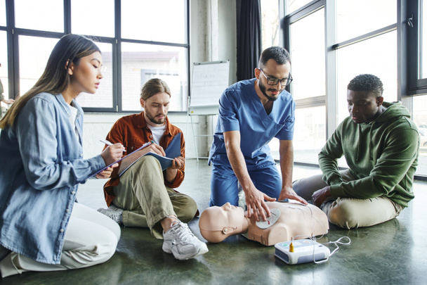 Herz-Wiederbelebung Fähigkeiten, Gesundheitshelfer Anwendung Defibrillator-Pads auf CPR-Schaufensterpuppe in der Nähe von jungen vielfältigen und multiethnischen Gruppe, Gesundheitsfürsorge und lebensrettende Techniken Konzept - Foto, Bild